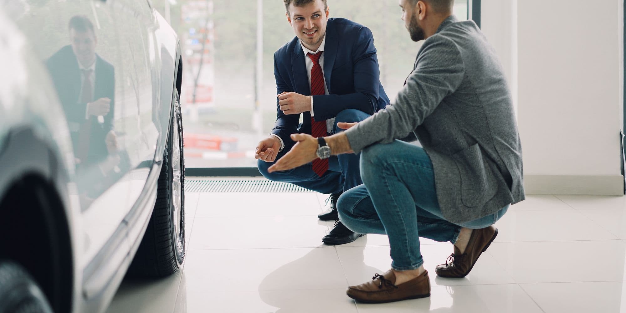 A salesperson and a customer crouched beside a car in a dealership showroom
