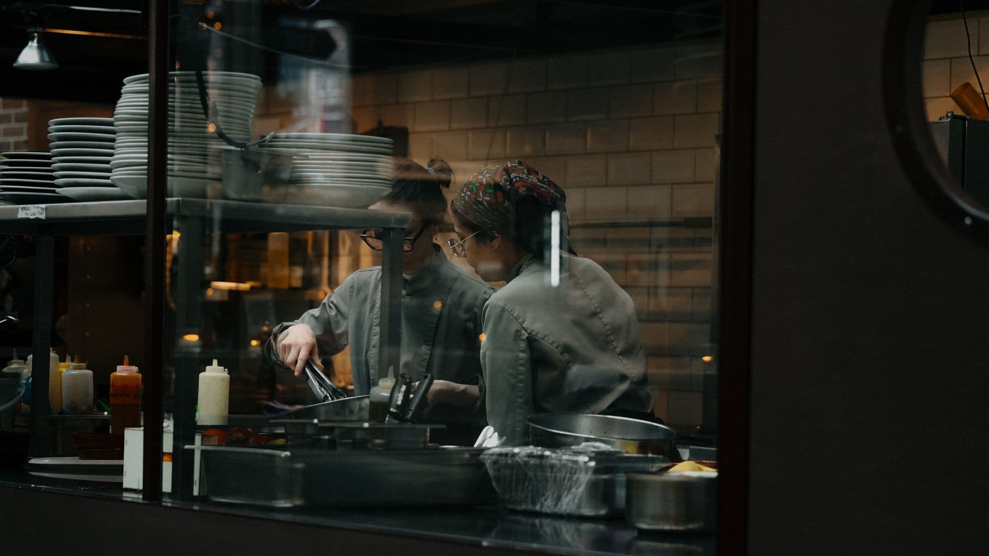 Two chefs working the pass in a restaurant kitchen seen through a window