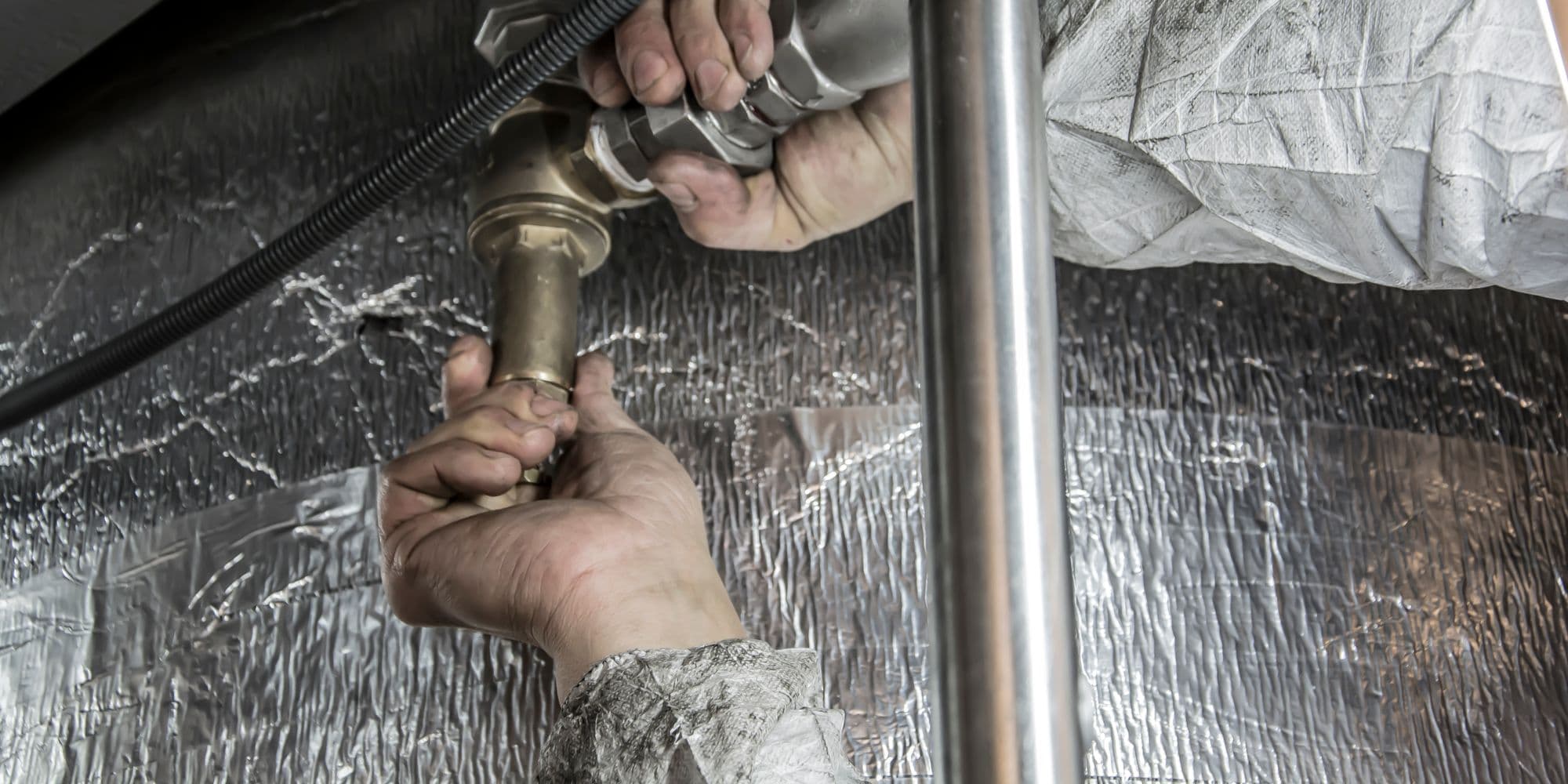 A pair of hands tightening a brass fitting on pipework under a unit