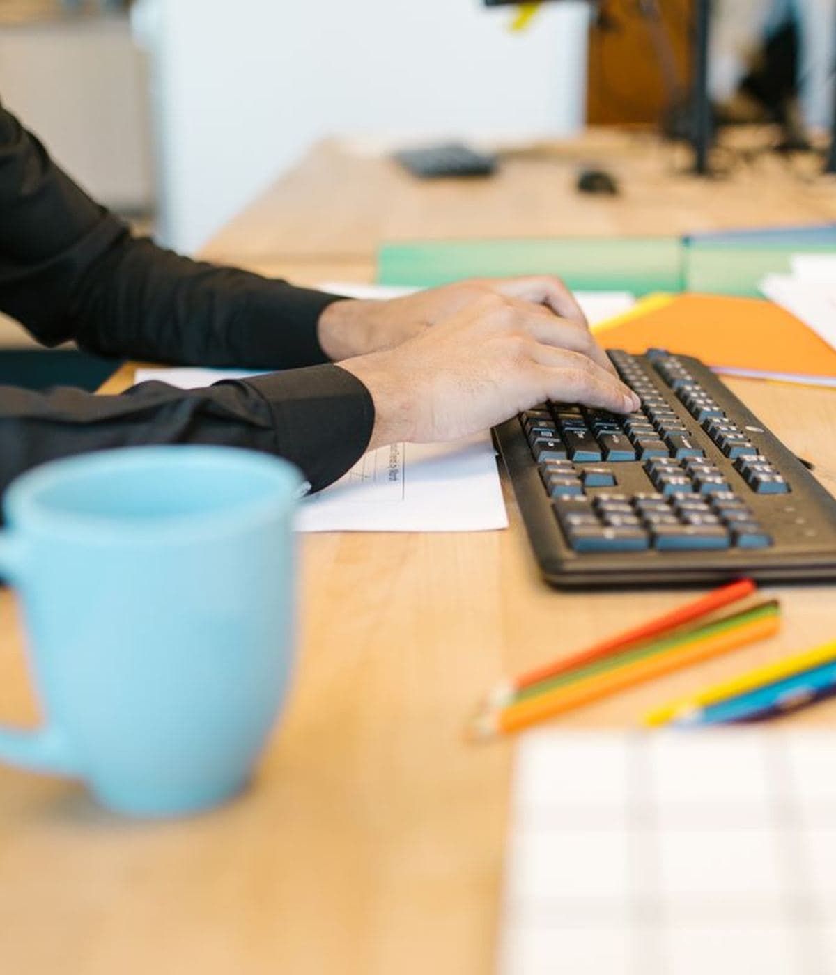 Hands at a keyboard on a warm wooden desk beside a blue mug and coloured pencils